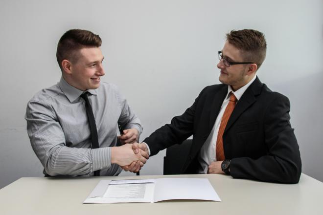 Two men in suits shake hands, sitting near some papers on a desk