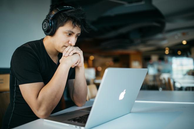 A student sits in a cafeteria looking intently at his laptop screen, hands clasped, with headphones on
